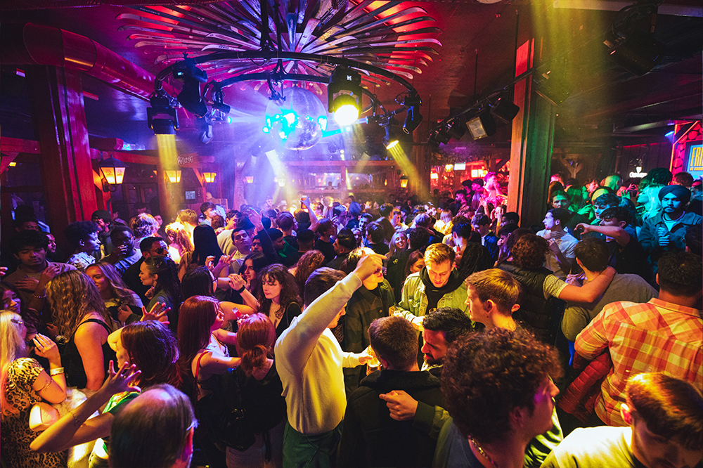 Crowd shot of people dancing and partying on a busy dance floor. Overhead colourful lights and a disco ball shine onto the crowd from above.